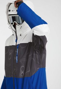 Persoon in een blauw, wit en zwart gepatternde skijas met capuchon, witte helm en reflecterende skibril, die één arm omhoog steekt.