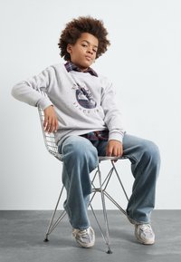 Felpa grigia con design grafico, sovrapposta a una camicia a quadri, indossata con jeans azzurri chiari e sneakers grigie, seduto su una sedia di filo.