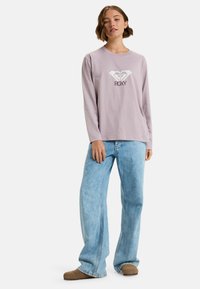 Ljuslila långärmad skjorta med vitt hjärta och "ROXY"-text; matchas med ljusblå vida jeans och bruna slip-on skor.