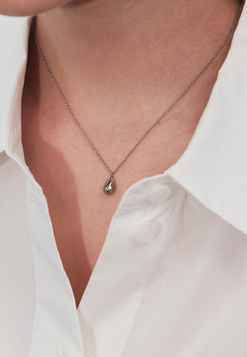 Strieborný prívesok v tvare kvapky na jemnom náhrdelníku, ležiaci na koži. Nosený s bielou košeľou s golierom a gombíkmi. Jednoduchý a elegantný dizajn.