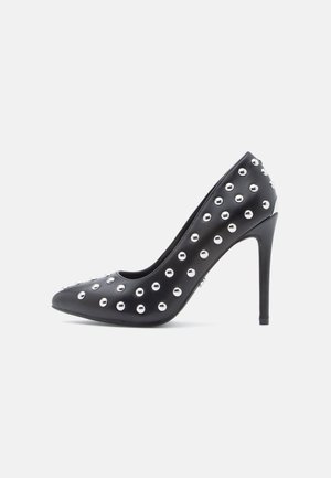 Black high-heel shoe with a pointed toe, made of smooth material, adorned with round silver studs across the upper and a slim stiletto heel.