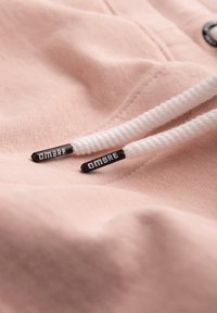 Witte trekkoorden met zwarte uiteinden met het label "OMBRE" op zacht roze stof, waarschijnlijk onderdeel van een hoodie of sweater.