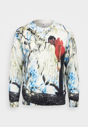 Pull avec un imprimé d'une personne en chemise rouge se penchant en avant au milieu de motifs abstraits bleu, noir et blanc évoquant des formes d'arbres.