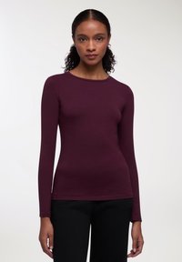 Długi rękaw w odcieniu głębokiego burgundu, wykonany z miękkiego materiału, z okrągłym dekoltem i dopasowanym krojem, zestawiony z czarnymi spodniami.