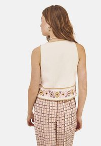 Top recortado sin mangas de color crema con una banda decorativa bordada en rosa y amarillo a lo largo del dobladillo, combinado con pantalones de cuadros en rosa y blanco.