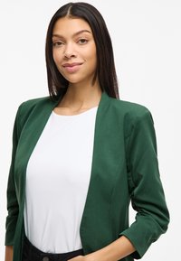 Grønn blazer med en jevn tekstur, åpen frontdesign og trekvartlange ermer, parret med en hvit topp og svarte bukser.