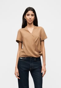 Γυναίκα με μακριά σκούρα μαλλιά που φοράει χρυσούς κρίκους στα σκουλαρίκια, καφέ μπλουζάκι με κοντά μανίκια και σκουρό μπλε τζιν, στέκεται μπροστά σε απλό λευκό φόντο.