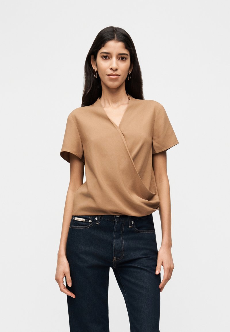 Γυναίκα με μακριά σκούρα μαλλιά που φοράει χρυσούς κρίκους στα σκουλαρίκια, καφέ μπλουζάκι με κοντά μανίκια και σκουρό μπλε τζιν, στέκεται μπροστά σε απλό λευκό φόντο.