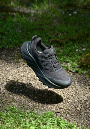 Mørkegrå New Balance løbesko ophængt ovenover en grussti med grønt græs og små blomster i baggrunden.