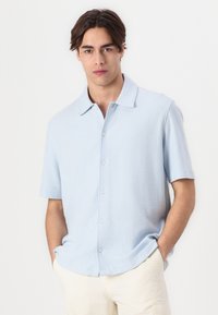 Camisa de manga corta azul claro hecha de tela texturizada, con un frente abotonado y cuello puntiagudo, combinada con pantalones de color crema.