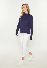 Marineblauer, gerippter Strickpullover mit hohem Kragen und verlängerten Ärmeln, kombiniert mit engen weißen Hosen und weißen Sneakern. Schlichtes, elegantes Design.