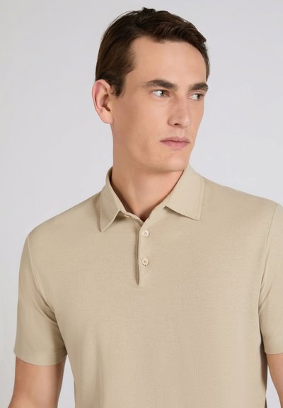 Man wearing a beige short-sleeve polo shirt, looking to the side against a plain light gray background.