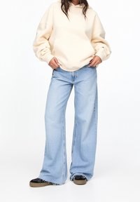 Světlý béžový mikina s přední kapsou, v kombinaci s širokými světlými modrými džínami. Džíny mají roztrhané lemy a kontrastní švy. Černé tenisky.