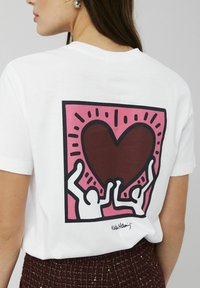 T-shirt en coton blanc avec un grand graphique au dos représentant un cœur brun, encadré par des contours roses et noirs avec des bras levés.
