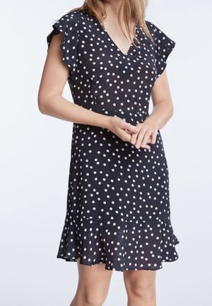 Robe noire à pois blancs, manches courtes et évasées, décolleté en V et ourlet volanté. Fabriquée en tissu léger et légèrement transparent.