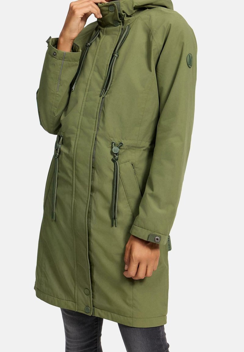 Long manteau parka vert avec zip et boutons-pression, cordons ajustables à la taille, poignets boutonnés et capuche, porté par une personne en jean foncé.