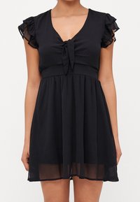 Černé krátké šaty s v-výst�řihem, detailem na zavazování vpředu a volánovými rukávy. Vyrobené z lehkého materiálu s splývavou sukní a přiléhavým pasem.