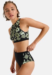 Mladá žena upravujúca vysoké bikini nohavičky s čiernym a zeleným kvetinovým vzorom, ktoré ladia s crop topom, na jednofarebnom pozadí.