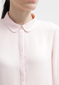 Blouse rose clair avec col rond, fermeture à boutons et texture lisse et douce. Présente un design classique et ajusté avec une légère brillance.