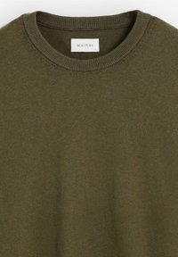 Suéter de punto verde oliva con cuello redondo y puños acanalados, que presenta un sutil logo bordado en el pecho. Textura suave.