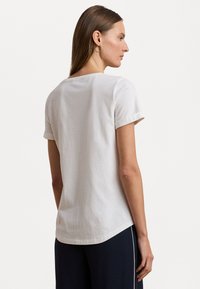 Balta medvilnė marškinėliai su apvaliu iškirpimu, trumpomis susukamomis rankovėmis ir šiek tiek kreivu apvadu, demonstruojami modelio iš nugaros.