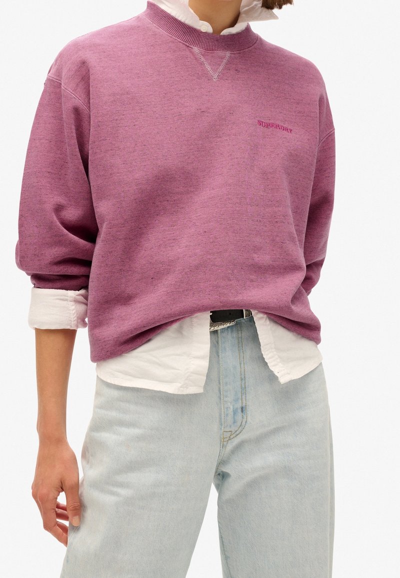 Personne portant un sweat-shirt col rond rose par-dessus une chemise blanche à boutons rentrée dans un jean bleu clair.