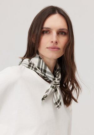 Kobieta o brązowych włosach nosi biały top i czarno-białą kratkowaną chustę zawiązaną na szyi na jednolitym tle.
