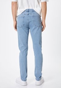 Jeans de mezclilla azul claro, de corte ajustado, con un diseño estándar de cinco bolsillos, canesú en la parte trasera y detalles de costura sutiles. Zapatillas blancas mostradas debajo.