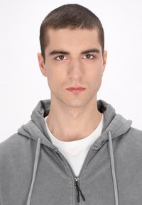 Szara bluza z kapturem o miękkiej fakturze, z zamkiem błyskawicznym z przodu i sznurkiem. Nosi ją na białej koszulce z okrągłym dekoltem. Model pozuje z poważnym wyrazem twarzy na jednolitym tle.