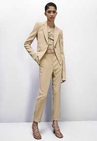 Completo beige su misura con blazer accorciato e pantaloni abbinati, abbinato a un top con motivo camouflage e sandali con tacco e cinghie.