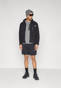 Svart fleecejakke over en grå skjorte, svarte shorts, gråstrikket lue og solbriller. Høye svarte sneakers og svarte sokker fullfører looken.