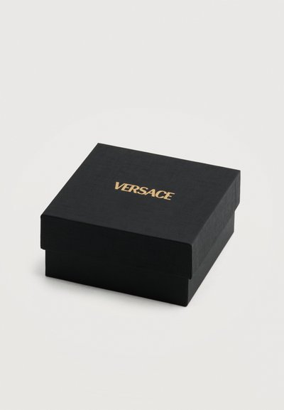 Μαύρο τετράγωνο κουτί Versace με αφαιρούμενο καπάκι, που φέρει χρυσό λογότυπο στο κέντρο της κορυφής, τοποθετημένο σε απλό ανοιχτόχρωμο φόντο.