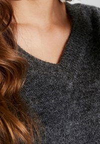 Maglione grigio scuro in lana con scollo a V e una superficie morbida e strutturata, parzialmente visibile con dei lunghi capelli castani mossi nelle vicinanze.
