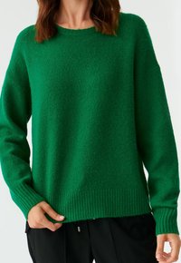 Femme portant un pull en maille vert à manches longues et un pantalon noir, tenant l'ourlet du pull avec la main gauche.