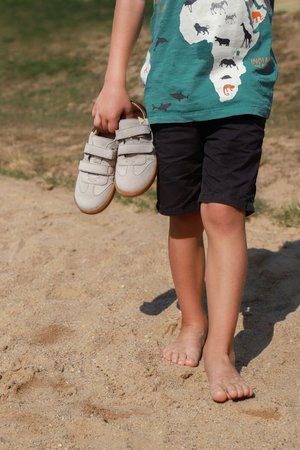 Child in animal-print green shirt and black shorts holds light gray shoes, standing barefoot on sandy ground outdoors.