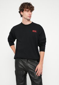 Melna kokvilnas garroku T-krekls ar sarkanu "DIESEL" logotipu kreisajā krūtīs. Kopā ar melnām ādas biksēm.