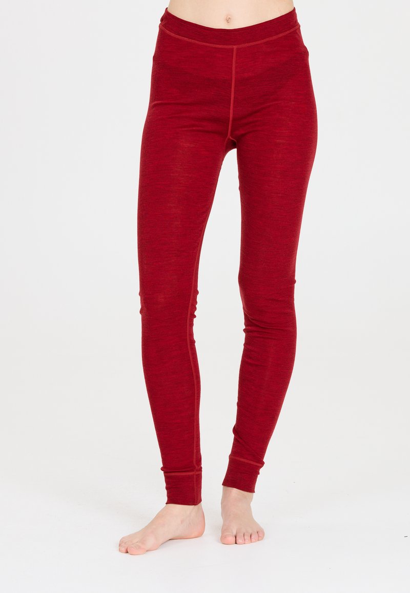 Persoon in strakke rode legging die blootsvoets staat tegen een effen witte achtergrond.