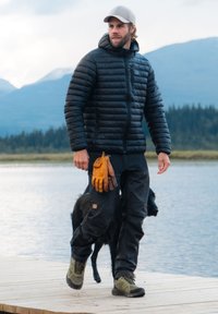 Zwarte pufferjack, zwarte broek, gele handschoenen en olijfgroene schoenen. Wandelen naast een zwarte hond bij een meer met bergen op de achtergrond.