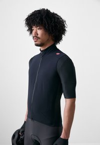 Uomo con capelli ricci che indossa una maglia da ciclismo nera e guanti, tiene un casco nero, volto a sinistra su uno sfondo semplice.