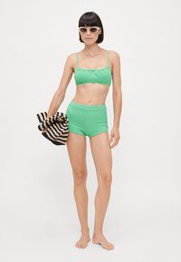 Top bikini verde a coste e pantaloni alti, abbinati a una borsa a righe nera e beige. Modella in piedi a piedi nudi, indossa occhiali da sole.