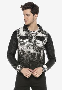 Schwarze Leder- und weiße Stoffjacke mit einem aufwendigen schwarzen Muster, zwei Brusttaschen und dekorativen Nieten entlang der Vorderseite.