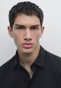 Portrait of a person with short dark hair and fair skin, wearing a black shirt. Features include a straight nose and full lips.