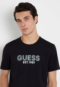Camiseta de algodón negra con cuello redondo que presenta "GUESS EST. 1981" en letras a rayas azul y blanca en el pecho.