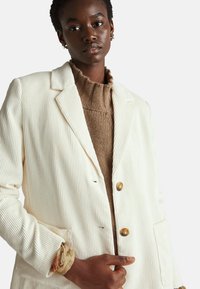 Krémový kordový blazer s ribkovou texturou, dvěma zlatými knoflíky a předními kapsami. Nositý přes hnědý pletený svetr.