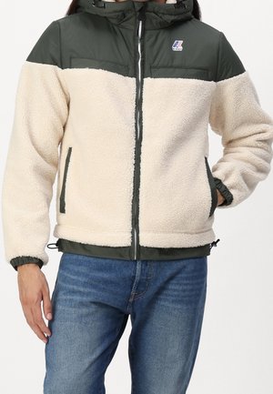 Homme portant une veste polaire bicolore verte et crème avec une fermeture éclair à l'avant et des poches latérales, associée à un jean bleu.
