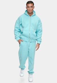 Completo di felpa blu chiaro con cappuccio e pantaloni della tuta abbinati, caratterizzato da una chiusura con zip, tasca a marsupio e polsini a coste. Sneakers bianche completano il look.