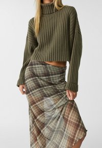 Olijfgroen geribbelde coltrui met verlaagde schouders, gecombineerd met een doorschijnende maxi-jurk in een ruitpatroon met aardetinten van bruin en groen.