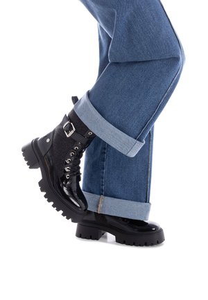 Botas negras con cordones y suela gruesa, hechas de materiales brillantes y texturizados. Llevadas con jeans azules arremangados que revelan puños en contraste.