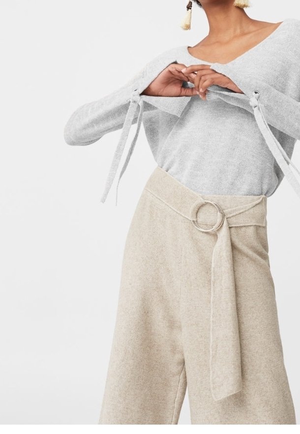 Femme portant un haut gris clair à manches longues avec découpes aux épaules et un pantalon beige taille haute avec une ceinture en tissu et une boucle métallique ronde.