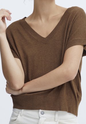 Persoon die een bruine, korte mouw, V-hals trui draagt en witte broek, met één arm gekruist en de andere hand dicht bij het gezicht opgeheven.